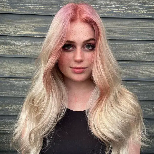 una mujer con una camisa negra sin mangas tiene el pelo rubio con raíces de color rosa pastel